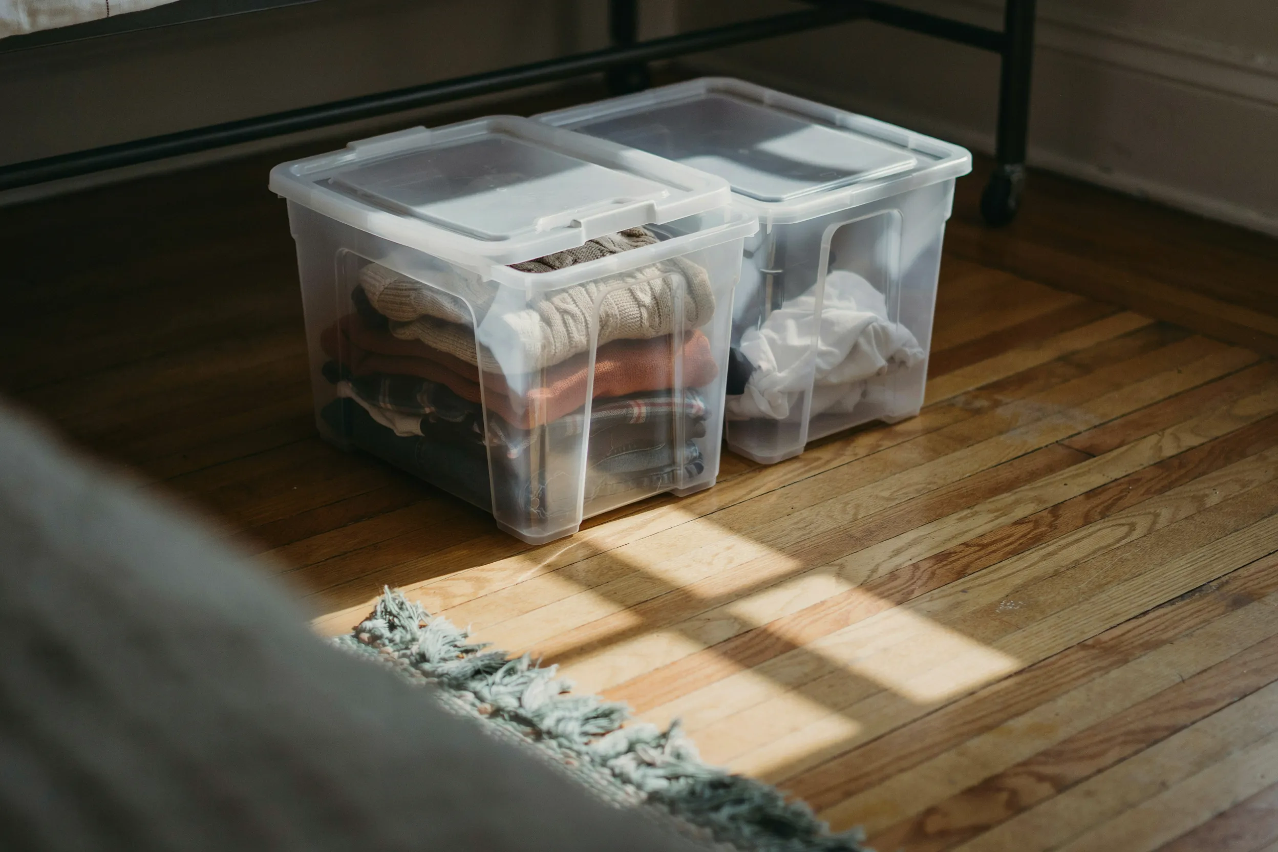 Two clear plastic storage bins with folded clothes inside, partially slid out from underneath a dorm bed frame