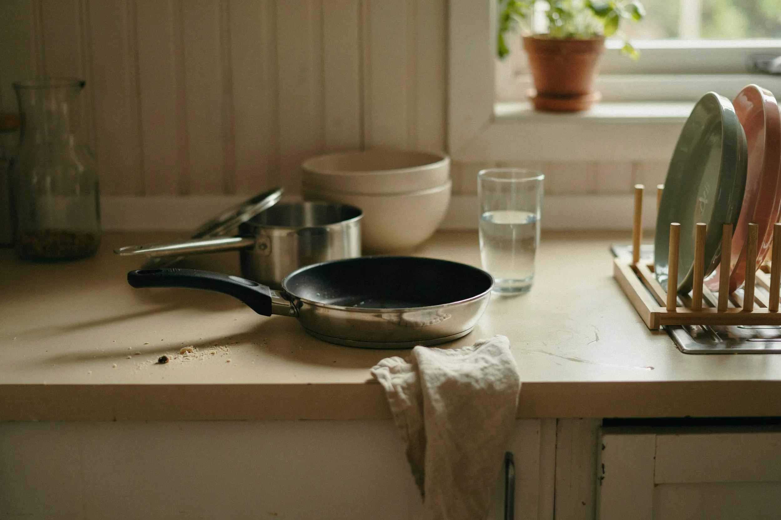 First apartment kitchen counter with a nonstick skillet, a medium pot, a stack of mixing bowls, and a wooden dish rack drying two plates