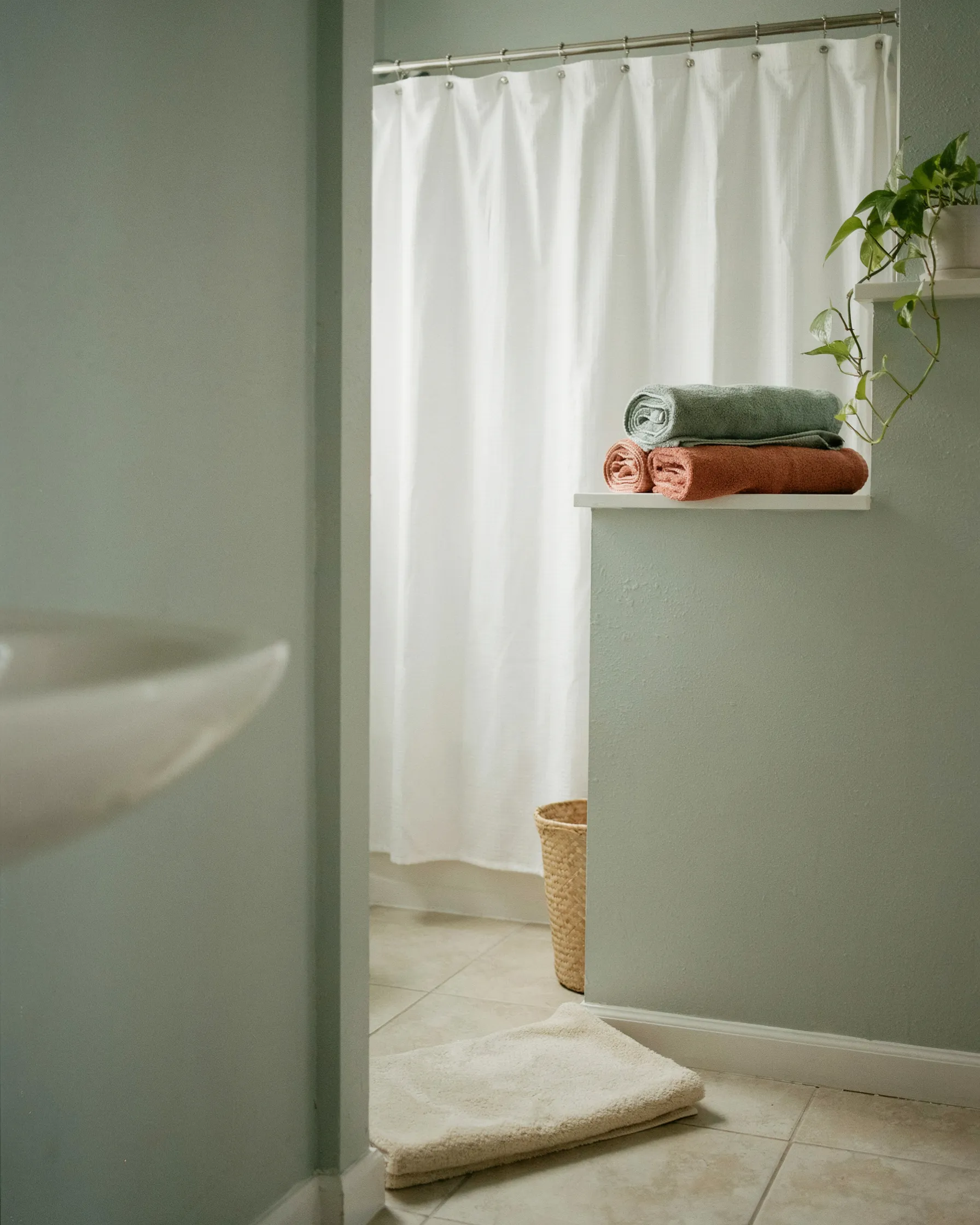 Small rental bathroom with a white shower curtain, rolled sage and terracotta towels on a shelf, and a folded bath mat on tile flooring