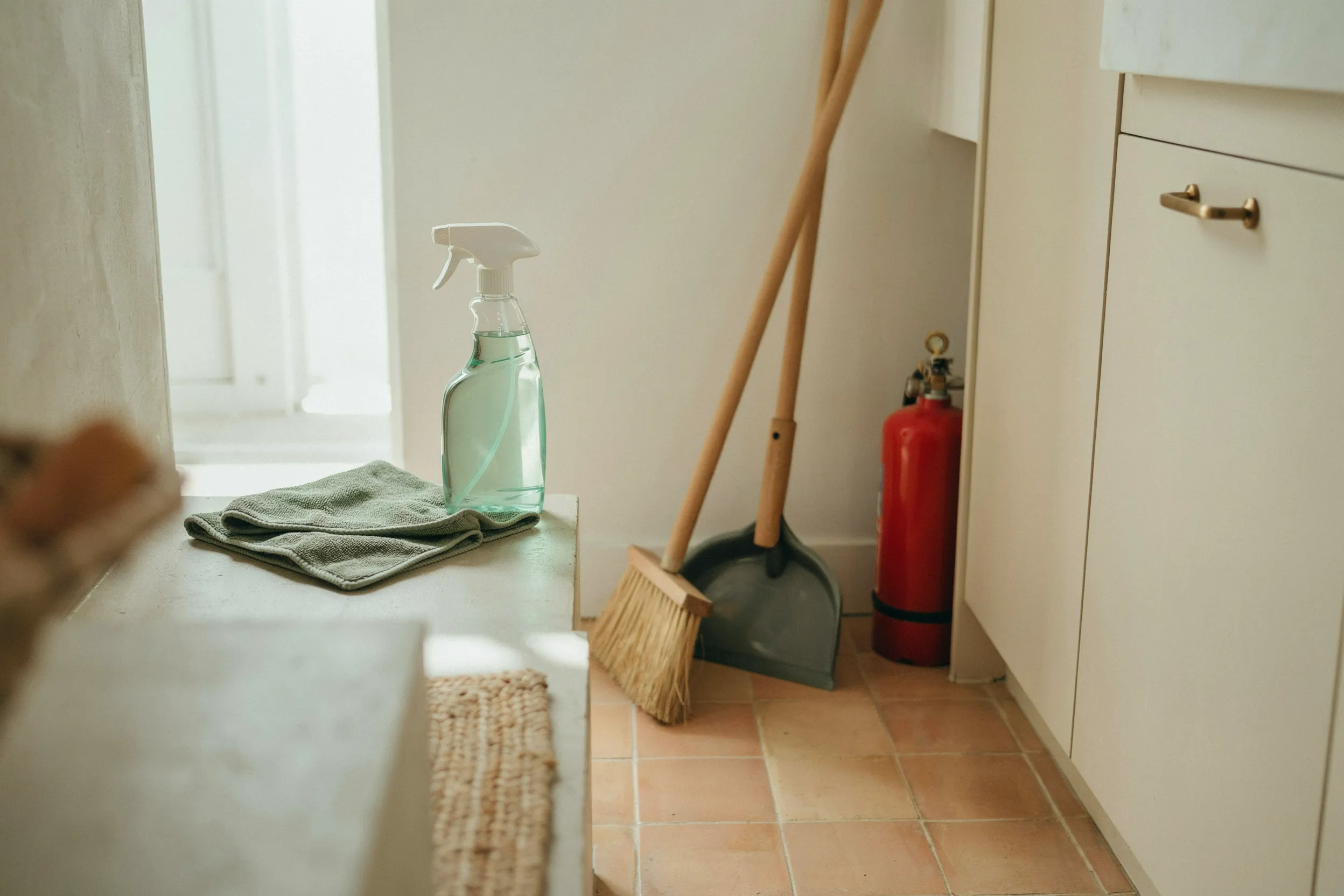 Apartment utility corner with a spray bottle of all-purpose cleaner, a microfiber cloth, a broom and dustpan, and a small fire extinguisher