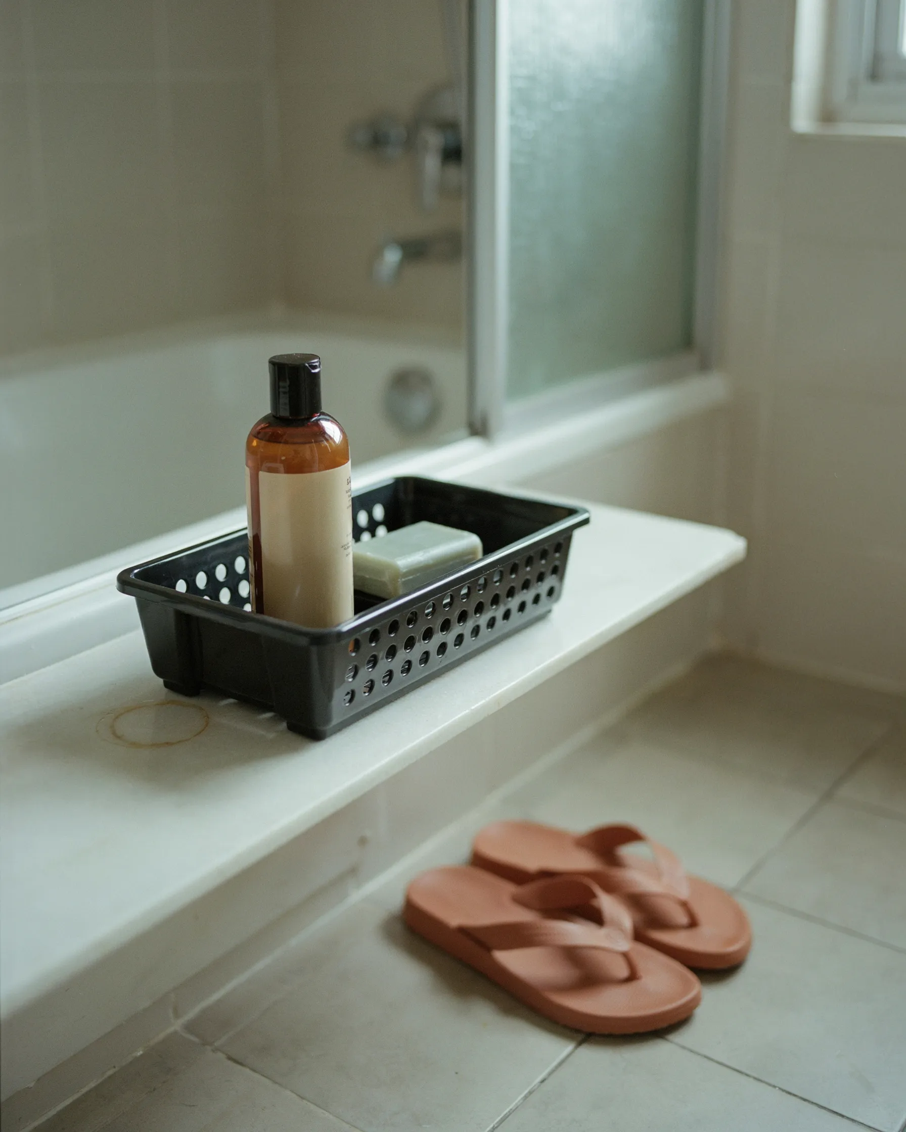 Shower caddy with drainage holes holding shampoo and soap, with a pair of terracotta flip-flops on the bathroom floor nearby